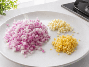 Finely chopped onion, minced garlic, and grated ginger prepared on a white plate in a bright modern kitchen with natural daylight.