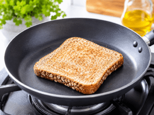 Bright realistic cooking step image of whole grain bread lightly warming inside a black nonstick frying pan on a gas stove in a modern kitchen.