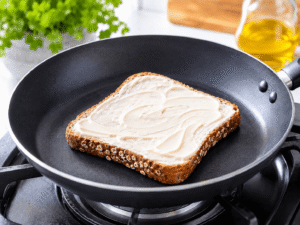 Close-up cooking image of mayonnaise evenly spread over toasted whole grain bread inside a black frying pan with bright natural kitchen lighting.
