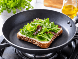Ultra realistic food photography of fresh mixed salad greens layered over mayonnaise on toasted whole grain bread inside a frying pan in a bright kitchen.