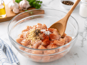 Raw chicken breast pieces in a glass bowl mixed with minced garlic, paprika, salt, and black pepper for chicken meal prep bowl recipe.
