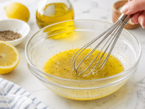Mixing olive oil, lemon juice, garlic powder, salt, and black pepper in a glass bowl to make a simple homemade dressing