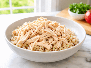 Cooked quinoa topped with shredded chicken breast in a white serving bowl on a marble countertop, prepared as the base for a high-protein chicken egg white bowl.
