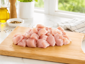Fresh raw chicken breast cut into small bite-size pieces on a wooden cutting board in a bright modern kitchen with natural daylight.