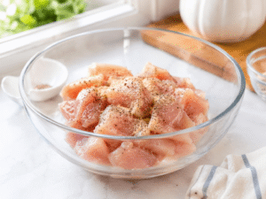 Raw chicken breast pieces in a glass bowl seasoned with salt, black pepper, and garlic powder on a bright kitchen counter.