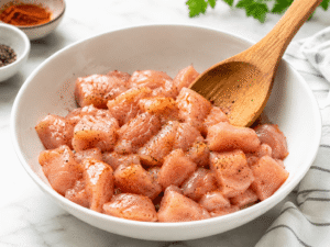 Raw chicken breast cut into bite-sized pieces and seasoned with salt, black pepper, and paprika in a white mixing bowl.
