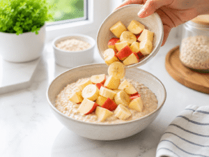 Fresh banana slices and apple chunks being added on top of a bowl of creamy oatmeal for a healthy breakfast.