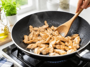 Thinly sliced chicken breast cooking in olive oil inside a black nonstick wok until lightly golden and fully cooked.