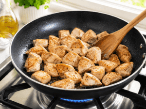 Chicken pieces cooking in a black skillet until golden brown while being stirred with a wooden spoon over a gas stove.