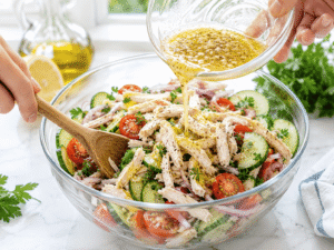 Lemon garlic dressing made with olive oil, lemon juice, garlic, salt, and black pepper being poured over chicken, cucumber, cherry tomatoes, red onion, and parsley before tossing.