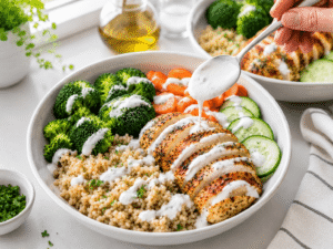 Creamy yogurt dressing being drizzled over a healthy chicken bowl filled with chicken, rice, broccoli, carrots, and cucumber.